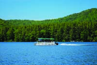 Squam Lake Cruise pontoon boat moving to the left across the water with tree covered hills in the background.